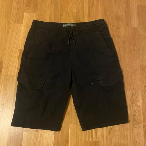 Svarta cargo shorts från Denim Co. - Svarta cargo shorts från Denim Co. med elastisk midja och snörning. Klassisk cargomodell med stora fickor på sidorna och avslappnad passform. Perfekta för sommardagar när du vill ha både stil och funktion.