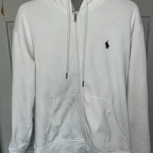Vit hoodie från Polo Ralph Lauren - Säljer en fin men ganska använd vit hoodie från Polo Ralph Lauren med dragkedja och justerbar huva. Det finns fläckar på några ställen men dom syns knappt när du har på dig den, jag är 191 cm och den passar mig bra. 