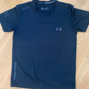 Svart tränings-tshirt Under Armour - Svart kortärmad t-shirt från Under Armour med diskret logga på bröstet och tryck på ärmen. Tillverkad i lätt och ventilerande polyester, perfekt för löpning eller gym. Fitted passform och rund hals. Snygg och sportig stil.