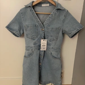 Kort jeansklänning med knappar - Snygg ljusblå jeansklänning från Zara med kort ärm, klassisk krage och knäppning framtill. Klänningen har fyra fickor, två på bröstet och två framtill nertill. Aldrig använd, prislapp och allt kvar! 