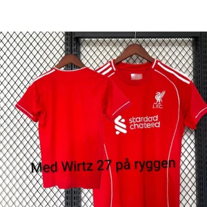 Liverpool röd fotbollströja Wirtz 27 addidas märket är mä fast de inte syns  - Röd Liverpool fotbollströja med vita detaljer och trycket 'Wirtz 27' på ryggen. Klassisk design med klubbmärke och sponsorlogga framtill, vita ränder på axlarna och smala vita linjer längs sidorna. Kortärmad och tillverkad i lätt, sportigt material. Helt oanvänd och med primer League-märke på axeln fast de inte syns och addidas märket fast de inte syns. Och du ska hest tvätta de för hand så att inte trycket försvinner 