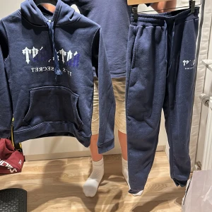 Trapstar sett (Mörkblå hoodie och byxor från Trapstar) - Säljer en mörkblå hoodie och byxor från Trapstar med vit och lila tryckt text på bröstet. Tröjan har huva med snörning, magficka och långa ärmar. Perfekt för dig som gillar streetwear och vill ha en chill look. 