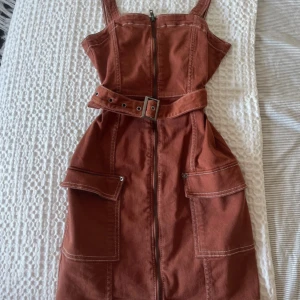 Rostbrun kort klänning H&M Divided - Rostbrun kort jeansklänning från H&M med breda axelband, bälte och fickor framtill. Klänningen har dragkedja hela vägen fram. Endast använd 1 gång och är så gott som ny! Passar xs-s.