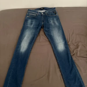 Replay Grover Hyperflex jeans blå - Snygga Replay Grover Hyperflex jeans i klassisk blå tvätt med slitningar på låren och knäna. Jeansen har fem fickor, normal passform och är tillverkade i stretchigt jeansmaterial för extra komfort. Perfekta för dig som gillar en stilren och avslappnad look. Storlek 32/34