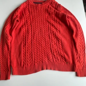 Orange stickad kofta Holly & Whyte - Säljer en röd stickad kofta från Holly & Whyte by Lindex i storlek M. Koftan har grovt flätstickat mönster, ribbade muddar och rund halsringning. Perfekt för lager-på-lager och ger en färgklick till din outfit.