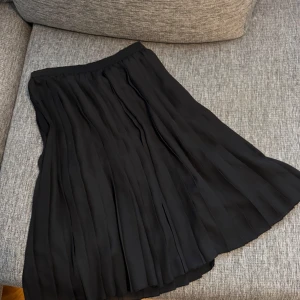 Svart plisserad kjol från Uniqlo - Säljer en svart plisserad kjol från H&M i storlek 38. Kjolen har en klassisk midja och är tillverkad i ett lätt, flowy polyestertyg som ger snyggt fall. Perfekt för dig som gillar stilrena och tidlösa plagg med lite extra rörelse.