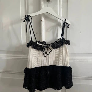 Stickad svartvit linnetopp med volang - Supersöt stickad linnetopp i svart och vitt med volangdetaljer och knytbara axelband. Toppen har en ribbad struktur upptill och en luftig, volangig nederdel. Perfekt för dig som gillar en lekfull och trendig stil.