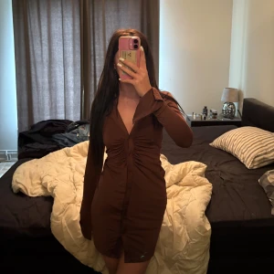 Brun kort skjortklänning med snörning - Säljer en brun kort skjortklänning med lång ärm och v-ringning. Klänningen har snygg snörning framtill som ger en cool rynkad effekt och knappar hela vägen ner. Perfekt för dig som gillar en tight och stilren look.