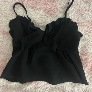 Svart linnetopp med volang och knyt - Supersöt svart linnetopp med smala axelband, volangdetaljer längs urringningen och knyt framtill. Toppen har en lätt utsvängd passform och smockad rygg för extra komfort. Perfekt att styla med kjol eller jeans för en trendig look.