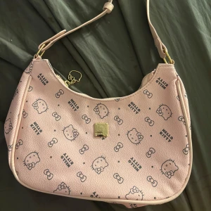 Hello Kitty rosa axelväska - Supergullig rosa axelväska från Hello Kitty med tryckta motiv av Hello Kitty och rosetter över hela väskan. Väskan har guldfärgade metalldetaljer, dragkedja med Hello Kitty-hänge och en fyrkantig logoplatta framtill. Tillverkad i syntetmaterial med lätt struktur.