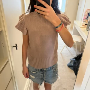 Beige stickad topp Holly & Whyte - Supersöt stickad topp i beige från Holly & Whyte. Modellen har ribbad hög hals och voluminösa, korta ärmar med snygg struktur. Perfekt till jeans eller kjol för en trendig look. Materialet är mjukt och skönt stickat.