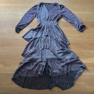 Glittrig långklänning från Maje - Säljer en glittrig långklänning från Maje i lila och silver. Klänningen har långa ärmar, v-ringning och flera lager volanger som ger en riktigt snygg siluett. Använd 2 ggr.