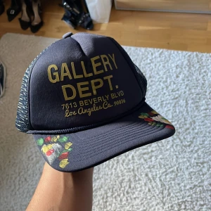 Svart truckerkeps Gallery Dept. - Snygg svart truckerkeps från Gallery Dept. med guldigt tryck framtill och handmålade färgdetaljer på skärmen. Kepsen har mesh på sidorna och justerbar baksida för perfekt passform. Perfekt statement-piece för dig som vill sticka ut.
