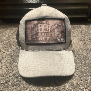 Grå keps från Sayless med stadsmotiv - Snygg grå keps från Sayless med svart mesh på sidorna och baksidan. Framtill finns en stor patch med ett coolt stadsmotiv i svartvitt. Kepsen har böjd skärm och justerbar passform. Perfekt för dig som vill sticka ut med en unik accessoar.