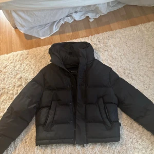 Trapstar Puffer Decoded Jacka - Säljer en svart pufferjacka från Trapstar med huva och broderad logga på ryggen. Jackan har dragkedja framtill, två stora fickor och en snygg patch på ärmen. Perfekt för kalla dagar och riktigt street i stilen.  litet hål vid armhålan, dm för bild