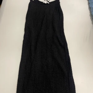 Svart ärmlös kortklänning - Säljer en svart ärmlös kortklänning med strukturerat tyg. Klänningen har enkel och stilren design med smala axelband och rak passform. Perfekt för dig som gillar minimalistisk stil och vill ha något bekvämt men snyggt.