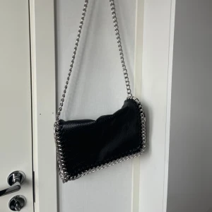 Svart axelväska med kedjedetaljer Tiamo -  Svart axelväska från Tiamo i skinnimitation med grova silvriga kedjor längs kanterna och som axelrem. Väskan har dragkedja upptill, ytterfack med dragkedja och flera innerfack. Hela väskan är i mycket bra skick.