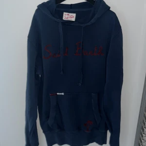Mörkblå hoodie från MC2 Saint Barth - Mörkblå hoodie från MC2 Saint Barth med röd broderad text på bröstet. Tröjan har huva med snörning, känguruficka och långa ärmar. Storlek S