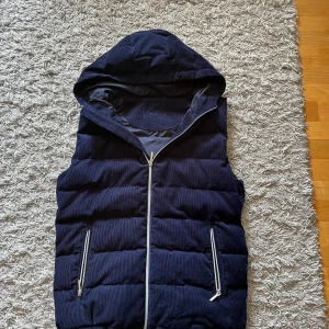 Marinblå manchester dunväst Vendiera - Snygg marinblå dunväst från Vendiera i manchester med huva och silvriga dragkedjor. Västen har quiltad design och två fickor framtill. Perfekt för lager-på-lager och ger en cool vibe till din outfit.