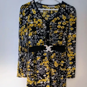 Blommig klänning med bälte från Bulaida - Färgglad långärmad klänning från Bulaida med svart botten och blommigt mönster i gult och grått. V-ringad med knappar framtill och ett brett svart bälte med glittrigt spänne i midjan. Perfekt för dig som gillar att sticka ut med coola detaljer.