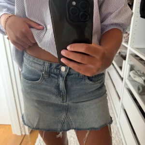 Blå jeanskjol med fransig kant - Säljer en snygg ljusblå jeanskjol i kort modell med fransig, rå kant nertill. Kjolen har klassiska fickor fram och bak, bälteshällor och dragkedja med knapp. Perfekt för en chill och trendig look.