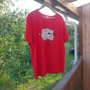 Röd t-shirt med fårtryck - Röd t-shirt i polyester med ett gulligt tecknat får på bröstet. Står att det är xxl men är som L Klassisk rund halsringning och korta ärmar. Perfekt för dig som gillar roliga och lekfulla motiv. Skön och avslappnad passform.