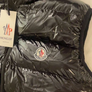 Svart dunväst från Moncler - Snygg svart dunväst från Moncler med glansig finish och klassisk logotyp på bröstet. Västen har dragkedja framtill och är perfekt för lager-på-lager-stil. Passar dig som vill ha en stilren och exklusiv look.