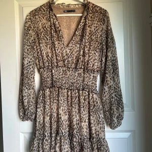 Leopardmönstrad kortklänning från Zara - Snygg kortklänning från Zara med leopardmönster i beige och bruna toner. Klänningen har v-ringning, smockad midja och långa ärmar med lätt puff. 