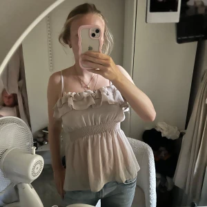 Beige volangtopp  - Supersöt beige linnetopp med volanger upptill och smockad midja. Tunna axelband och luftig passform som ger en romantisk vibe. Perfekt till jeans eller kjol för en chill look. 