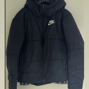 Svart pufferjacka från Nike S - Svart pufferjacka från Nike med huva och dragkedja framtill. Jackan har ett stilrent Nike-logga på bröstet och är quiltad för extra värme. Perfekt för kalla dagar och har en clean streetstyle-vibe.