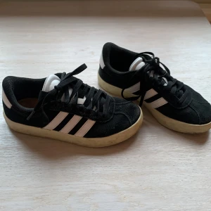 Adidas svarta sneakers - Klassiska Adidas sneakers i svart mocka med vita detaljer och de ikoniska tre ränderna på sidorna. Låg modell med snörning och platt sula. 