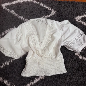 Vit blus med spets från Pimkie - Supersöt vit blus från Pimkie med bred smockad midja, puffiga korta ärmar och v-ringning dekorerad med spetsdetaljer. Blusen har även spets längs ärmarna och en luftig, romantisk känsla. Perfekt för dig som gillar vintage vibes och detaljer.