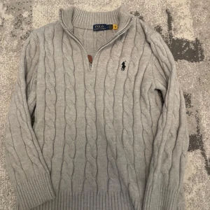 Grå kabelstickad tröja Polo Ralph Lauren - Kabelstickad grå tröja från Polo Ralph Lauren med halv dragkedja och broderad logga på bröstet. Tröjan har hög krage, långa ärmar och ribbade muddar. Perfekt för lager-på-lager och chill höst- eller vinterstil. Passar till storlek M också tro mig. Pris kan diskuteras vid snabb affär!