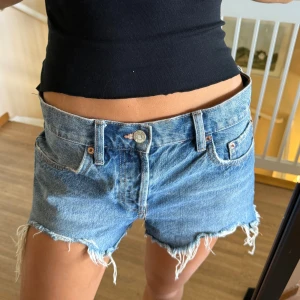Blå jeansshorts med fransig kant - Snygga blå jeansshorts från Levi's med låg midja och rå, fransig kant. Klassisk femficksmodell med knapp och dragkedja framtill. Perfekta för sommaren och ger en avslappnad vibe till din outfit.