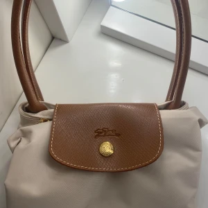 Beige Longchamp le pilage  - Beige och brun Longchamp le pilage väska. Väskan har guldknapp med logga och klassisk broderad häst på locket. Perfekt för skolan, jobbet eller varje dag! Helt ny, aldrig använd innan! Storlek M! 