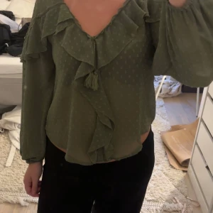 Olivgrön blus med volanger och knyt - Säljer en olivgrön blus med diskret prickigt mönster, djup v-ringning och volangdetaljer längs halsringning och framtill. Blusen har långa, luftiga ärmar och knyt med tofsar vid halsen. 