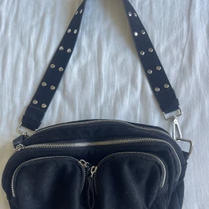 Svart axelväska i mocka med nitar - Cool svart axelväska i mocka med bred axelrem dekorerad med silvriga nitar. Väskan har flera fack med dragkedjor och silvriga metalldetaljer. Perfekt för dig som vill ha en edgy och praktisk väska till vardags💕