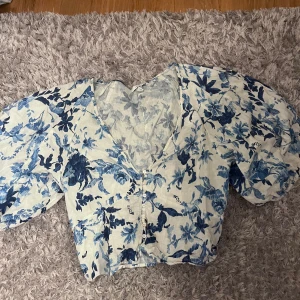 Blommig linnetopp från H&M, blå/vit - Superfin blommig linnetopp från H&M i blått och vitt. Toppen har puffiga korta ärmar, v-ringning och knappar framtill. Perfekt för sommarens alla tillfällen 