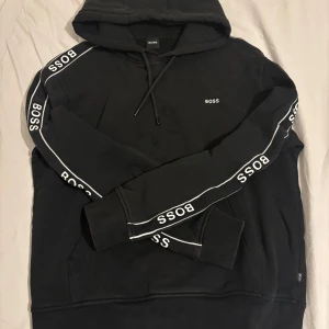 Svart hoodie från BOSS med logga - Svart hoodie från BOSS med vit logga på bröstet och längs ärmarna. Tröjan har huva med snörning och ribbade muddar. Klassisk och stilren design med sportig känsla, perfekt för dig som gillar streetwear.