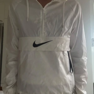 Vit vindjacka från Nike XS - Snygg vit vindjacka från Nike i storlek XS. Jackan har huva, halv dragkedja, elastiska muddar och stora svarta Nike-loggor fram och bak. Tillverkad i glansig polyester med praktiska fickor och justerbar nederkant. Perfekt för en sportig och clean look.