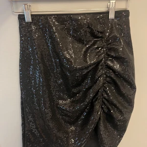 Svart glittrig kjol från H&M - Supersnygg svart glittrig kjol från H&M med paljetter över hela och snygg rynkdetalj på ena sidan. Kjolen är kort och har en figurnära passform som verkligen sticker ut. Perfekt för dig som vill ha något extra till din outfit! Några av paljetterna har ramlat av men inget man tänker på, se bild  4 & 5