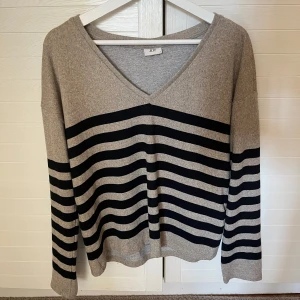 Randig v-ringad tröja från JDY - Beige och svart/blå randig tröja från JDY med v-ringning och lång ärm. Tröjan har en loose passform och är tillverkad i mjukt material, perfekt för lager-på-lager. Snygg till jeans eller kjol för en chill look.