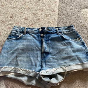 Blå jeansshorts - Snygga blå jeansshorts från NA-KD med hög midja och råa, fransiga benslut. Klassisk femficksmodell med silverfärgade knappar och slitna detaljer. Perfekta för varma dagar och enkel att matcha med allt.
