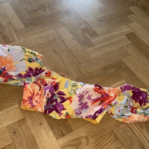 Blommig offshoulder topp i gult - Färgglad offshoulder topp med stora puffärmar och blommigt mönster i lila, orange, rött och vitt på gul botten. Toppen har croppad passform, smockdetaljer och knytband i ryggen. Perfekt för sommarens alla äventyr!