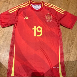 Spaniens landslagströja Lamine Yamal - Röd fotbollströja från Adidas för Spaniens landslag med nummer 19 och Lamine Yamal på ryggen. Tröjan har gula detaljer, tre ränder på axlarna och landslagets emblem på bröstet. Tillverkad i lätt funktionsmaterial med korta ärmar och modern passform.