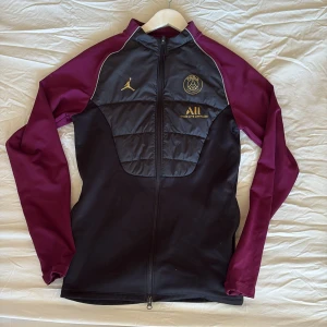 PSG x Jordan svart och vinröd jacka XS - Snygg PSG x Jordan jacka i svart med vinröda ärmar och guldiga detaljer. Jackan har hel dragkedja, quiltad front och loggor på bröstet. Materialmix med polyester och syntet, perfekt för fotboll eller chill. Långärmad och sportig vibe.