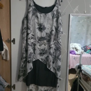 Blommig långklänning i lager-på-lager - Snygg långklänning med lager-på-lager-effekt i svart och grått. Klänningen har ett blommigt mönster och är ärmlös med rund halsringning. Perfekt för dig som gillar en flowy och unik stil.