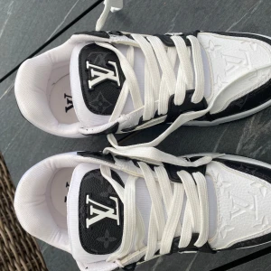 Louis Vuitton svartvita sneakers - Snygga svartvita Louis Vuitton sneakers med tydlig LV-logga på plösen och präglat monogrammönster i lädret. Skorna har vita snören, robust sula och coola detaljer med både präglad och tryckt logga. Perfekta för dig som vill sticka ut med lyxig streetstyle.