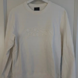 Vit sweatshirt från Oscar Jacobson - Vit sweatshirt från Oscar Jacobson med broderad logga framtill. Klassisk rund halsringning och långa ärmar. Tröjan är i mjuk bomull och har ribbade muddar vid ärmslut och nederkant. Perfekt för en clean och stilren look.