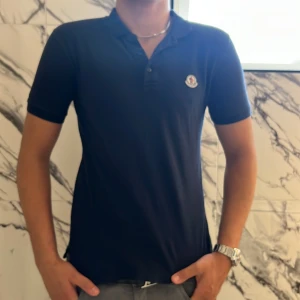 Mörkblå Moncler pikétröja S - Snygg mörkblå pikétröja från Moncler med klassisk krage, tre knappar och broderad logga på bröstet. T-shirten är kortärmad och i mjuk bomull, perfekt för en clean och stilren look.